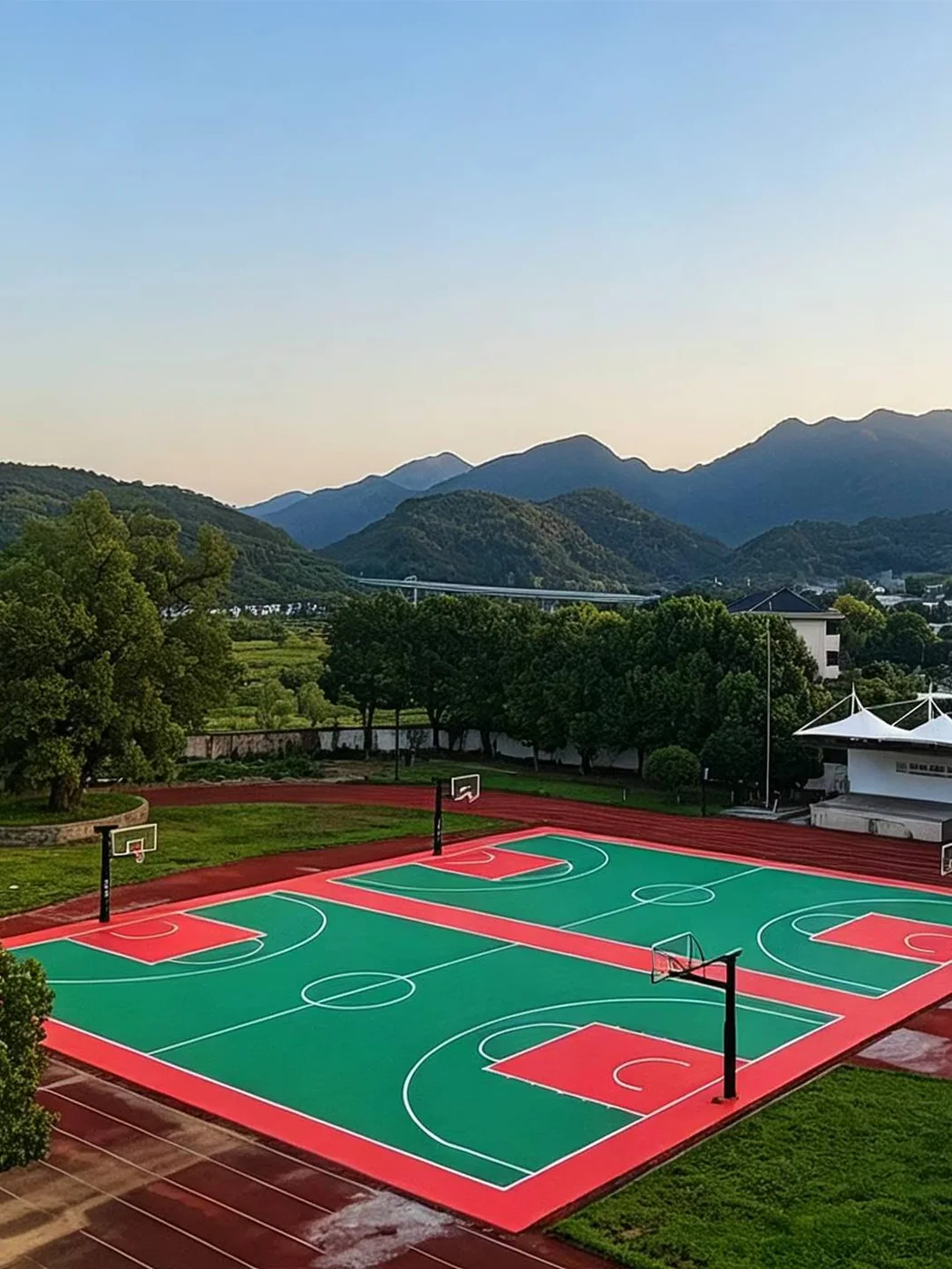 Two red and green outdoor basketball courts beside a running track and wooded hills